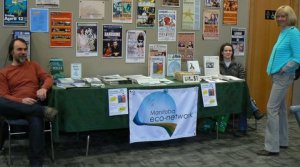 Manitoba Eco-Network display at the Living Lakes Concert image of a table with information pamphlets and 2 people sitting at either end